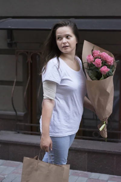 A girl walks from the store down the street with a paper bag and a bouquet of roses.