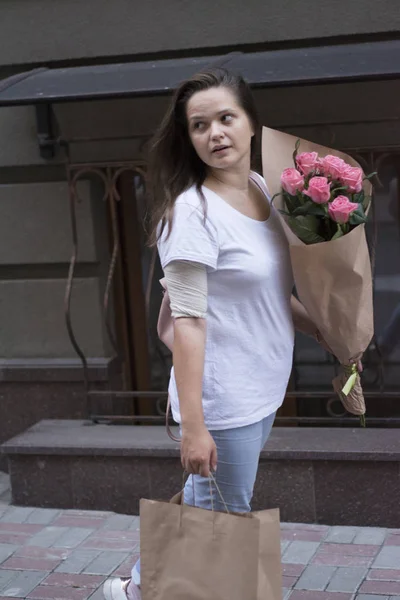 A girl walks from the store down the street with a paper bag and a bouquet of roses.