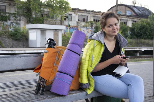 The girl sits with a backpack and camping equipment at the station.