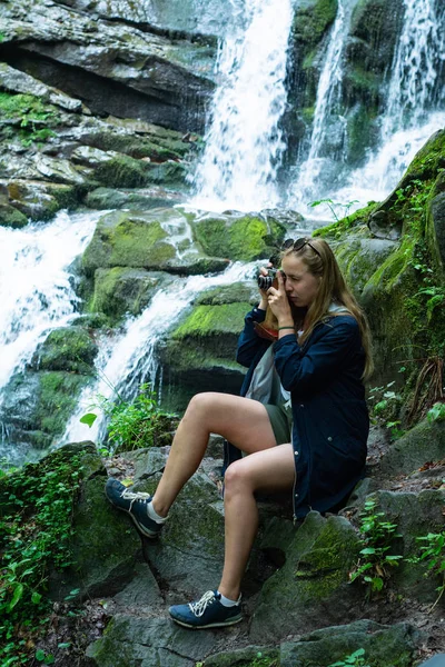 The girl is sitting with a camera on the background of a waterfall.