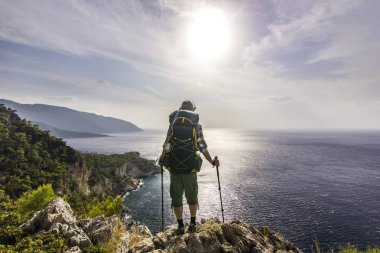 tourist with backpack standing on a cliff in mountains near mediterranean sea clipart