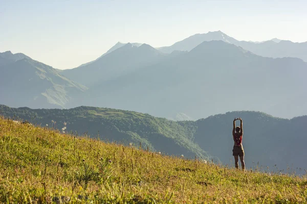 Meisje in jurk met handen omhoog staande op gras in zonsondergang bergen — Stockfoto