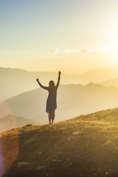 Дівчина в одязі з руками вгору, стоячи на траві в горах заходу сонця — стокове фото