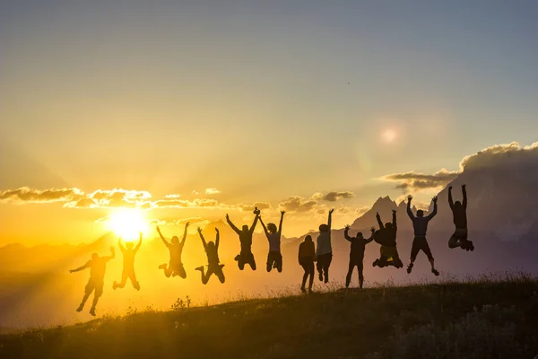 Csoport emberek kezeket fel ugrott a fű naplemente-hegység — Stock Fotó