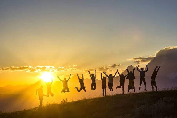 Group of people with hands up jumping on grass in sunset mountains — Stock Photo, Image