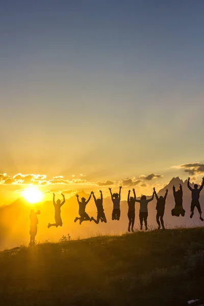 Group of people with hands up jumping on grass in sunset mountains — Stock Photo, Image