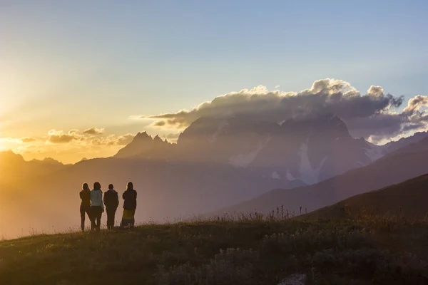 Grupo de pessoas com as mãos para cima em pé na grama em montanhas pôr do sol — Fotografia de Stock