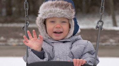 Salıncakta sallanan gülümseyen çocuk. Sevimli küçük çocuk (yürümeye başlayan çocuk, iki yaşında) kürk döşeme ile şapka ve güzelliğini. Mutlu bir çocuk. Kış, soğuk hava, açık
