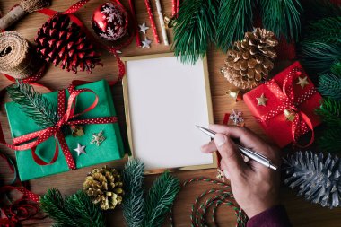 İnsan eliyle tebrik kartı yazan mutlu noel kavramları hediye paketi ve ahşap masa arkaplanındaki süs eşyası. Kış mevsimi etkinliği fikirleri.