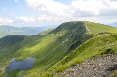 Parlak mavi gökyüzünün altındaki yemyeşil dağ yamaçlarının geniş panoramik manzarası, arazinin ortasına yerleşmiş sakin bir Dogyaska gölü içeriyor. Svydovets, Karpatlar, Ukrayna