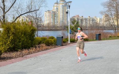 Atletik giyimli bir kadın nehir kenarındaki yolda koşuyor. Şehir zemininde koşuyor. Formunu, kararlılığını ve doğal ortamdaki kentsel yaşam tarzını tasvir ediyor.