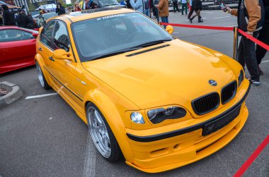 Açık turuncu BMW E46, vücut kiti ve Hamann markasıyla özelleştirilmiş, açık hava araba fuarında park halinde. Arabanın siyah ızgarası ve özel tekerlekleri var.
