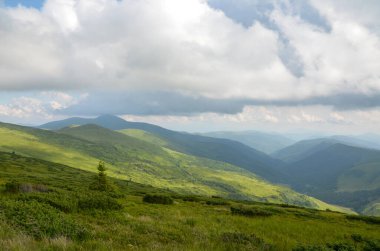 Parçalı bulutlu bir gökyüzünün altında geniş yemyeşil dağların bulunduğu bir dağ vadisinin manzarası doğanın sakin güzelliğini ve sakin atmosferini gözler önüne seriyor. Karpat Dağları, Ukrayna