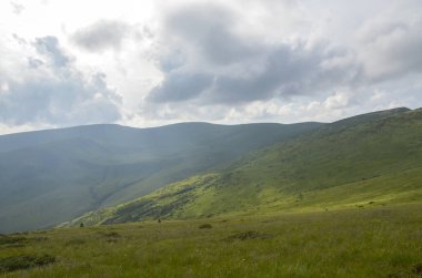 Dinamik parçalı bulutlu gökyüzü altında yuvarlanan yeşil dağ sırasının sakin manzarası. Sakin sahne doğanın güzelliğini ve dokunulmamış açık alanların huzurlu ortamını gösteriyor. Svydovets Karpatlar