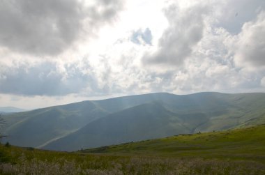 Dinamik parçalı bulutlu gökyüzü altında yuvarlanan yeşil dağ sırasının sakin manzarası. Sakin sahne doğanın güzelliğini ve dokunulmamış açık alanların huzurlu ortamını gösteriyor. Svydovets Karpatlar