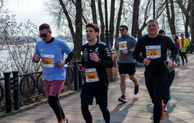 Bir maraton etkinliğinde nehir kenarında koşan bir grup sporcu, kararlılık, sağlık ve toplumsal katılımı sergiliyor