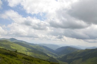Bereketli yeşil tepeler dramatik bulutlu gökyüzü ve güneş ışığı altında uzanıyor, dinginlik ve doğal güzelliği çağrıştırıyor. Resimli manzara huzurlu manzaranın barışçıl cazibesini somutlaştırıyor. Svydovets, Karpatlar