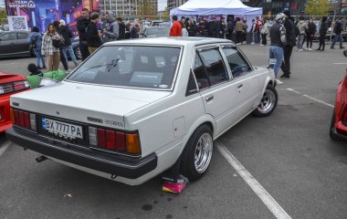 Parlak krom tekerlekleri olan klasik beyaz Toyota Carina sedan 'ın arka tarafında. Aracın eski usul Japon tasarımı var. Kutu şekli ve yatay stop lambaları var.