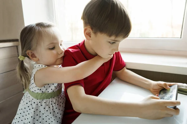 Hermana y hermano están sentados en la cocina y jugando con el teléfono —  Fotos de Stock