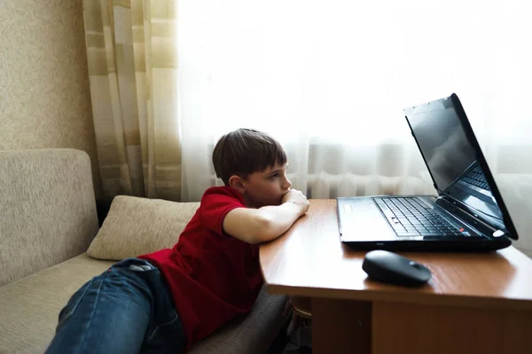 El chico yace en la sala de estar en el sofá y ve una película en una computadora portátil . —  Fotos de Stock