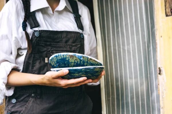 A woman in a white shirt and overalls is standing with ceramic dishes — Stock Photo, Image