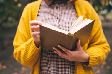 Bir kadın parkta yürüyor ve eski bir kitap okuyor. Turuncu hardal renginde moda sonbahar kıyafetleri.