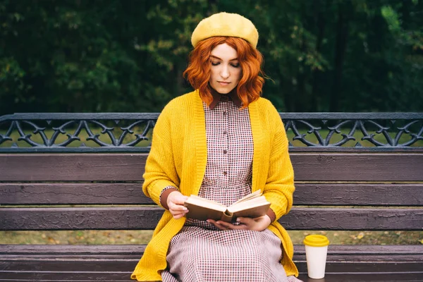 Kıvırcık saçlı güzel kızıl saçlı bir kız bir bankta oturuyor ve kitap okuyor. Yakınlarda bir bambu fincanı ve sıcak kahve duruyor. Turuncu hardal renginde moda sonbahar kıyafetleri.