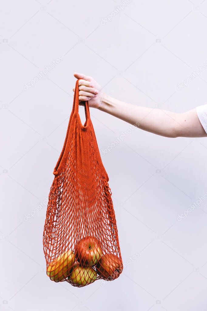 Female hand holds a red cotton shopping bag with oranges and apples on a background of white wall. The concept of naturalness and life with zero waste