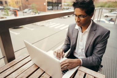 Genç ve yakışıklı Hintli bir adam elinde dizüstü bilgisayarla yazlık kafede oturuyor.