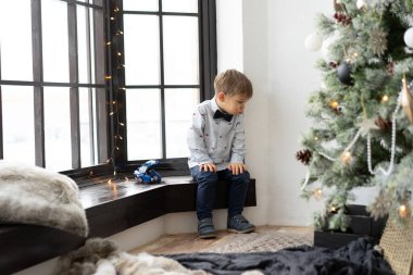  Küçük çocuk Noel ağacının yanındaki pencere pervazında oturuyor. 