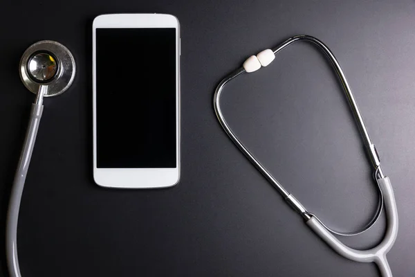 Medical stethoscope tool over the surface of a mobile smart phone, composition isolated over the black background.Checking healthcare security concept.