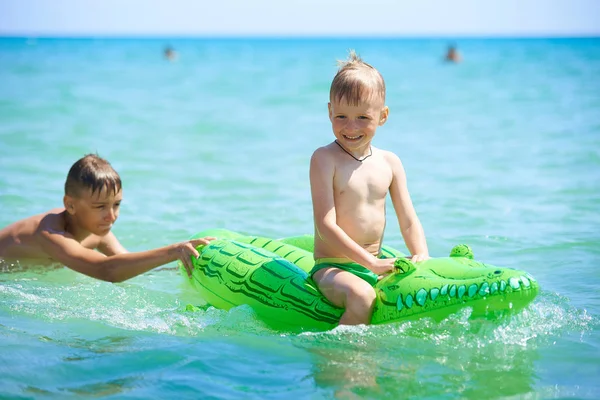 Older brother teenager rolls younger brother into the sea on a toy inflatable crocodile