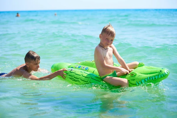 Older brother teenager rolls younger brother into the sea on a toy inflatable crocodile