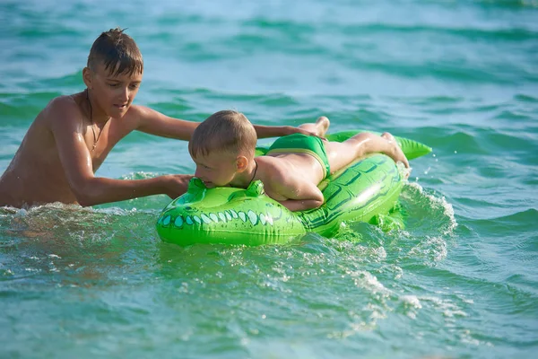 Older brother teenager rolls younger brother into the sea on a toy inflatable crocodile