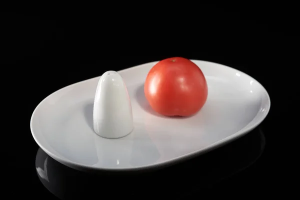 Un tomate vegetal y salero en un plato blanco, sobre un fondo negro — Foto de Stock