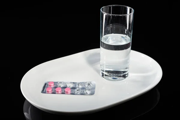 Copo de água e pílulas médicas em uma placa branca em um fundo preto . — Fotografia de Stock