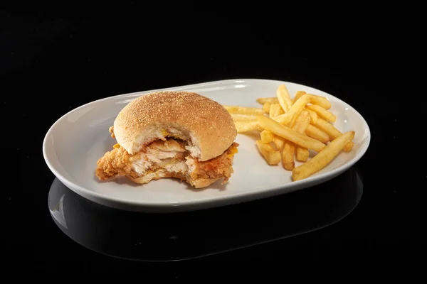 A fried chicken burger bitten on one side and a french fries on a white plate. The dish is on a sulfuric background.