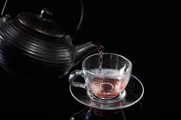 Chá quente vermelho é derramado a partir do bule em uma xícara de vidro em um fundo preto — Fotografia de Stock