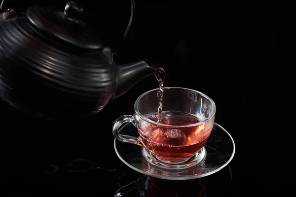 Té caliente rojo se vierte de la tetera en una taza de vidrio sobre un fondo negro — Foto de Stock