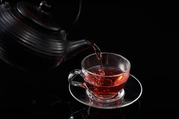 Té caliente rojo se vierte de la tetera en una taza de vidrio sobre un fondo negro — Foto de Stock