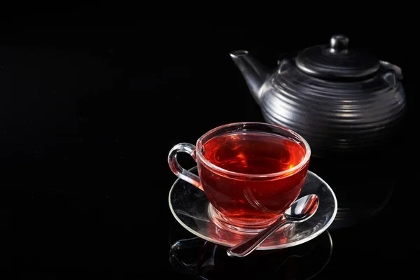 Glazen kopje met hete rode thee en theepot op een zwarte achtergrond. — Stockfoto
