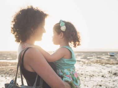 Küçük kız annesi gün batımı sırasında sahilde kollarında. Arka planda düşük gelgit nedeniyle ıslak kum görebilirsiniz. Yumuşak stiliyle Vintage filtre