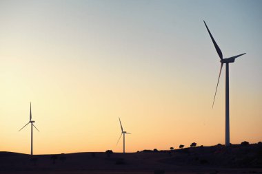 Üç yel değirmenleri rüzgar çiftliği yeşil enerji günbatımında üreten silüeti. Onlar bitkileri ve ağaçları ile çevrili bir kırsal ortamda, temiz ve berrak gökyüzü. Kopya alanı