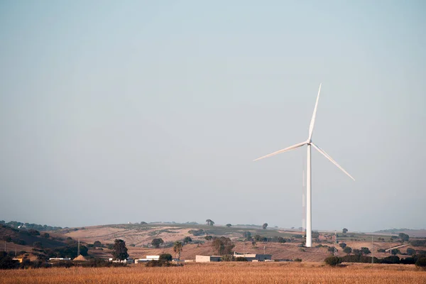A windmill produces clean energy in a wind farm. It is in a rural environment surrounded by crops and small hills, the sky is clean and clear. Copy space