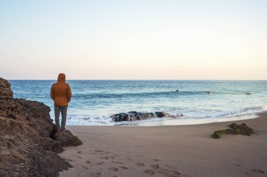 içinde birkaç adam sörf mavi ve turkuaz deniz kıyısında kayalar dikiz kahverengi bir ceket giymiş bir adam görünüyor, bu kış ve soğuk, ama açık gökyüzünde güneş parlıyor