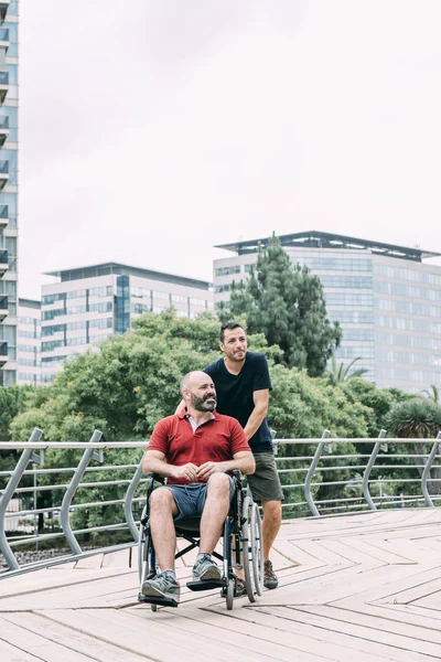 Man in wheelchair walking with his friend at city — Stock Photo, Image