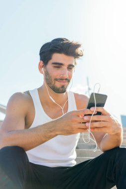 Kulaklıklı yakışıklı bir sporcunun dikey fotoğrafı müzik dinleyip telefonuna, teknoloji ve sağlıklı yaşam tarzına danışıyor.