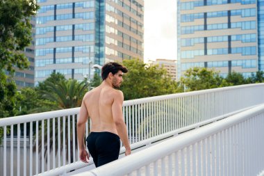 Spor adamı binaların önündeki köprüde üstsüz yürüyor. Şehir sporu ve sağlıklı yaşam tarzı kavramı.