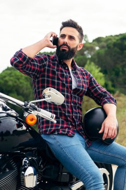 Kolunun altında kaskla motosiklet üstünde oturan yakışıklı sakallı bir adamın dikey fotoğrafı. Özgürlük kavramı ve motosikletçi yaşam tarzı.