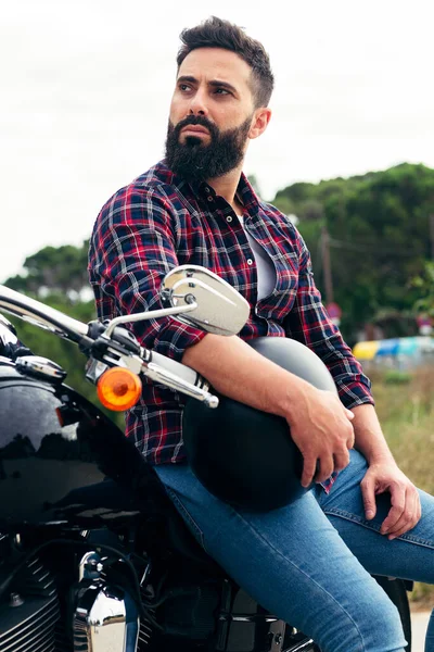 vertical photo of a handsome bearded man waiting on his motorbike with helmet under his arm, concept of freedom and biker lifestyle